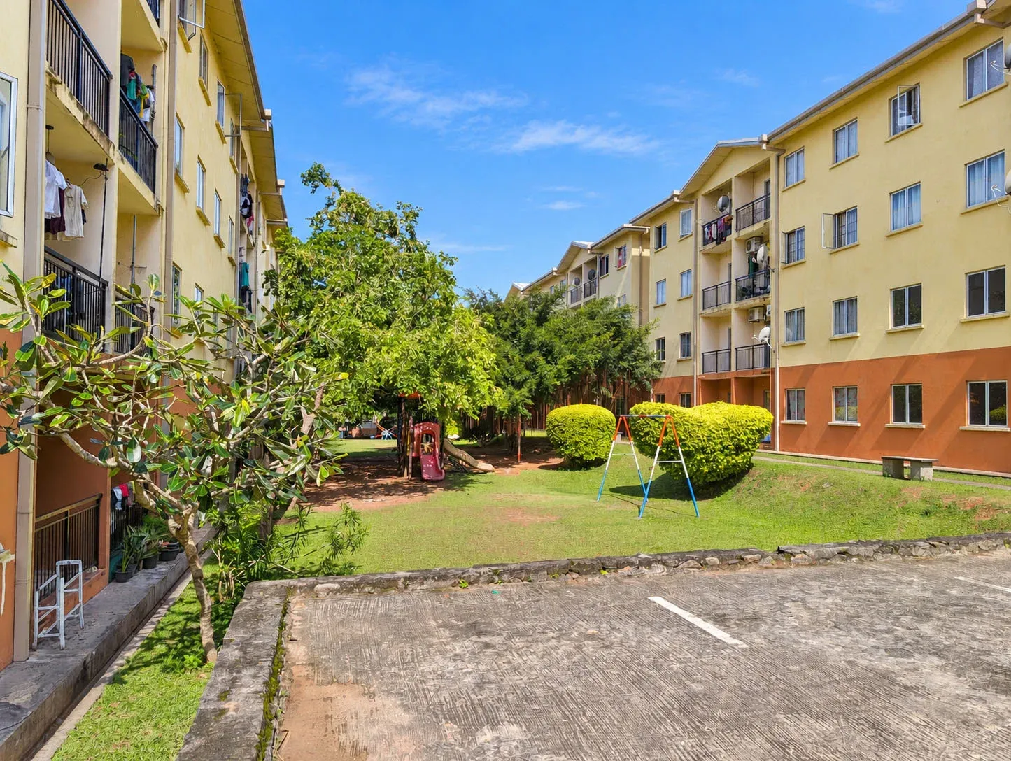 A shared children's play area within the apartment complex, with swings and slides in a grassy courtyard surrounded by trees. A nearby bench provides a place for parents to sit while children play.